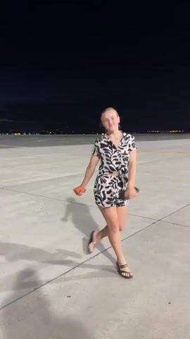 A young woman with blonde hair tied up in a bun is dancing on a concrete surface at night. She is wearing a black and white patterned shirt and shorts. She is holding a black object in her right hand and an orange object in her left hand. In the background, there are lights and a dark sky. She spins around and strikes poses while dancing.