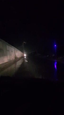 This media file captures a nighttime drive on a wet road in Paramankeni, India. The scene is dark, indicating it's likely late at night or early morning. Rain is evident from the wet surface of the road, which reflects the headlights of the vehicle the footage is being filmed from, as well as those of an oncoming vehicle. The oncoming vehicle's lights appear white and bright, creating a glare that momentarily obscures details of the road ahead.

To the left of the frame, a long, concrete barrier or wall runs along the side of the road. Its surface is textured and appears to be illuminated by the vehicle's headlights. The right side of the frame is mostly dark, with some faint lights visible in the distance, suggesting the presence of other vehicles or distant structures. There's a hint of purple and blue light reflecting on the wet road on the right side, possibly from a traffic signal or another light source.

The overall mood is one of solitary travel through challenging weather conditions. The focus is on the road ahead and the driving experience itself, with the rain and darkness creating a somewhat atmospheric and isolated feeling. No people are clearly visible, and no text is discernible within the footage. The activity is simply driving, with the focus on navigating the road under the given conditions. The perspective is from inside a vehicle, likely the driver's point of view, looking through the windshield.