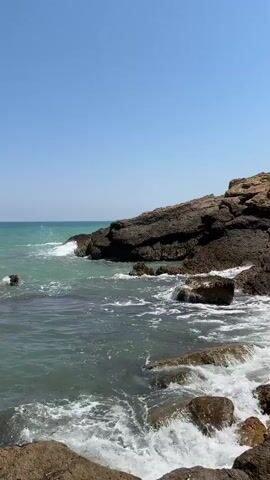 The video shows a rocky coastline with the ocean in the foreground and a clear blue sky in the background. Waves are crashing against the rocks, creating white foam. The water is a greenish-blue color. The rocks are dark brown and appear to be volcanic in origin. The sun is shining, casting shadows on the rocks. The scene is peaceful and serene.
