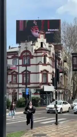 The video shows a street scene in what appears to be a city, possibly in Georgia based on the lettering on the billboard. The sky is partly cloudy, and the scene is captured during daylight. There's a large billboard above a building, displaying a portrait of a bearded man with a cross, adorned in clerical attire. The text on the billboard appears to be in Georgian. The building beneath it is a multi-story structure with red accents on its white facade, exhibiting a unique architectural style. Pedestrians can be seen crossing the street, with one woman in a black outfit prominently walking towards the camera. Several cars are moving in the street, including white sedans and a van. A street sign with digital information is also visible, partially obscured by the sun's glare, and it is surrounded by bare trees. The sidewalks are paved with interlocking bricks.