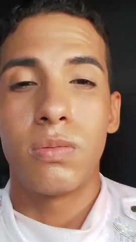 A man with dark curly hair and wet skin is shown in a close-up, selfie-style video. He is wearing a white shirt with some silvery, decorative elements on the shoulders. He looks down at first and then slowly looks up with a slight smile. The background is dark and blurry, possibly inside a vehicle. The man’s skin appears shiny and wet, as if he has just been sweating or gotten wet. He makes a slight pout with his lips. He appears to be talking but no audio is provided. He then smiles again and looks into the camera.