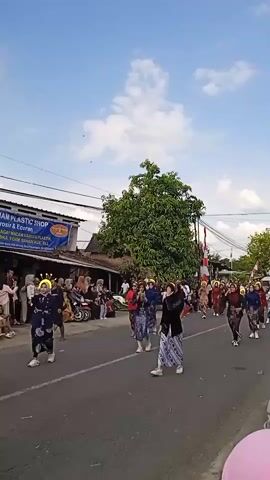 A crowd of people are participating in an outdoor parade on a sunny day. Most of them are wearing traditional batik clothing. Some people are dancing in the street, holding red scarves. On the left side of the road, spectators are sitting or standing. A plastic shop named "ARMAHAM PLASTIC SHOP" is visible in the background. A large green tree stands to the right of the shop. The sky is blue with scattered clouds.
