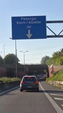 A car is driving on a highway toward a short tunnel under a bridge. The car is gray, and there are other cars ahead, also entering the tunnel. There are streetlights on either side of the highway, and trees are visible on the right side. The highway sign above the road points downward and reads Esch/Alzette A4.
