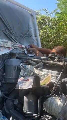 A person is working on the engine of a vehicle. The engine appears to be old or in disrepair, with visible parts and wiring. The person is wearing a bandana around their face, possibly for protection. They are using tools, likely to clean or maintain the engine components. The background shows foliage, suggesting an outdoor setting, possibly a yard or a workshop. The scene is brightly lit by natural sunlight.