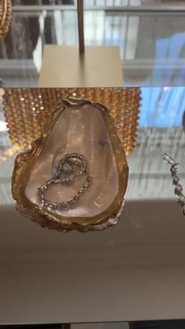 The video opens with a close-up view of a decorative oyster shell, its outer edges painted gold, resting on a highly reflective, clear surface. Inside the shell, a sparkling, silver-toned necklace or bracelet, adorned with numerous clear, round-cut stones, is intricately coiled.

As the camera slowly pans right, more jewelry comes into view. To the right of the oyster shell, a long, dangling earring or necklace segment is visible, featuring multiple rectangular, clear-cut stones set in a silver-toned metal. Further right, a pair of matching dangling earrings, each with several round, clear stones, hang vertically, with their reflections clearly visible on the polished surface below.

The background, consistently out of focus, reveals elements of a luxurious interior. Glimpses of a golden, intricately textured light fixture or chandelier are seen, casting warm, blurry reflections. Vertical lines of what appear to be crystal strands or beaded curtains also reflect in the background. A bright blue reflection, possibly from the sky or a large window, suggests ample lighting.

No people are visible in the frame. The scene portrays an elegant and high-end jewelry display, likely within a boutique or store in Beverly Hills, emphasizing the brilliance and design of the precious items. The smooth camera movement guides the viewer's attention across the gleaming, reflective objects. There is no visible text.