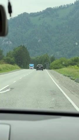 The video, filmed from the perspective of a driver inside a vehicle, shows a two-lane road with a truck and a car ahead. The road is bordered by lush green trees and a large, verdant mountain range in the background. The sky appears overcast. The camera is positioned behind the windshield, and the dashboard is visible in the foreground. The truck in the distance is blue and white with "OZON" branding. The car ahead is dark-colored. As the video progresses, the truck and car move further away, and other vehicles become visible further down the road. The scene is peaceful and conveys a sense of travel through a natural landscape.