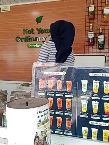 A person wearing a dark blue hijab and a striped long-sleeved shirt stands behind a counter. They are facing away from the camera. In front of the counter, there are several displays of drinks. To the left, there is a menu for "MENU MATCHA" showing pictures of matcha drinks. To the right, there are two larger menus: one titled "Tea Series" and another titled "Squash Series." Both menus display images of various drinks with their names and prices. There are also glass jars and bottles visible on the counter. A white cylindrical object with a slot on top, resembling a donation box, is placed prominently in the foreground. The background features a wooden wall with a green logo that reads "Not Your Ordinary Tea." The overall scene appears to be an outdoor beverage stand.