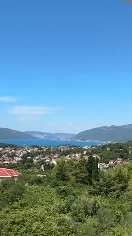 The video shows a wide shot of a coastal town with mountains in the background and a clear blue sky. An airplane is visible in the sky, flying diagonally across the frame and leaving a faint contrail behind it. The town appears to be built on a hillside overlooking the water, with buildings and greenery visible. The sea is a deep blue, and the mountains are covered in lush vegetation. The video is short, lasting only a few seconds.