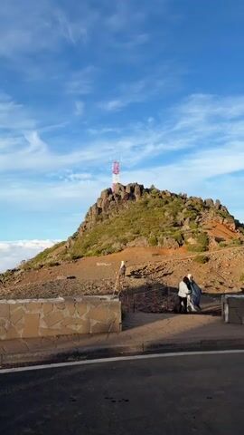 The video shows a scenic view from what appears to be a mountain top. A rocky slope with a communication tower on top is visible to the right of the frame. Below the slope is a stone wall barrier with a paved path. A person carrying equipment stands near the path. Behind the slope and barrier, the video captures a breathtaking view of mountains enveloped in a sea of clouds beneath a partly cloudy sky. The camera pans from the rocky slope and tower to the left, revealing more of the landscape with clouds and mountains.