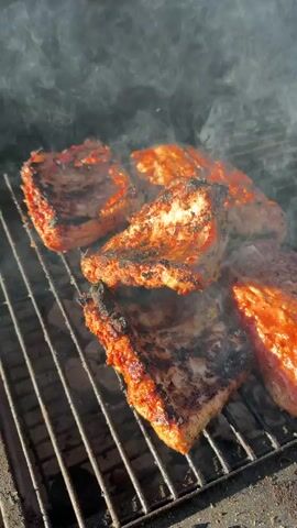 The video shows several racks of ribs being grilled. The ribs are covered in a reddish-brown sauce and are showing grill marks. Smoke is rising from the grill, indicating that the ribs are cooking. The grill grates are visible, and they appear to be made of metal. The background is blurred, but it seems to be an outdoor setting. The lighting suggests it is either morning or late afternoon.