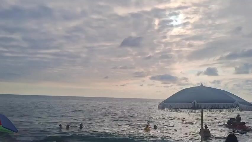 The video shows a beach scene with a cloudy sky. The sun is partially visible through the clouds. The ocean is calm with gentle waves. Several people are in the water, swimming and playing. There is a large blue umbrella with fringe on the right side of the frame, and a colorful umbrella on the left. A jet ski is visible in the water on the right. The video appears to be taken during daylight hours, possibly late afternoon or early evening.