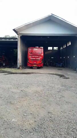 A red double-decker bus is parked inside an open-air garage. In the background, a person is visible in front of a red and black bus. Further back, there are several motorcycles parked. To the left of the garage, a yellow truck is parked. Inside the garage, a white car and a red car are visible. Another bus, black and yellow, is parked in the garage. Next to it, a yellow truck is parked. A person in a red shirt and blue jeans is standing near a truck. In the foreground, two men are standing near a truck covered with a blue tarp.