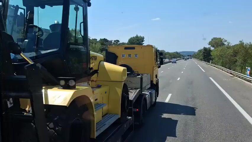 The video shows a Hyster forklift being transported on a flatbed truck. The forklift is yellow and appears to be large and heavy-duty. The truck is also yellow and is driving on a highway. The weather is sunny and clear. The video is shot from the perspective of someone on the truck, looking backward at the forklift. The forklift is secured to the truck with chains. The truck is driving in the right lane of the highway, and there are other vehicles on the road. The scenery outside the highway consists of trees and hills. The speed limit sign on the highway shows 110, indicating the truck is traveling on a highway with a speed limit of 110 km/h.