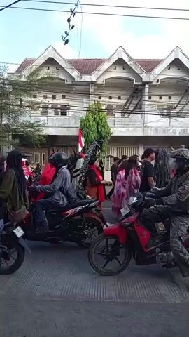A crowd of people are gathered on a street, with many riding motorcycles and scooters. Some individuals are wearing traditional clothing or costumes. The buildings in the background appear to be multi-story structures with peaked roofs. There are trees and electrical wires visible. The scene suggests a local event or parade.