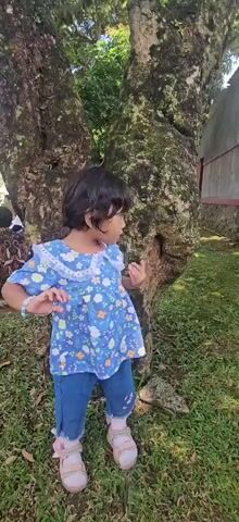 A young girl with dark hair is standing in a grassy area next to a large tree. She is wearing a blue dress with a white floral collar and blue jeans. Her arms are raised and her hands are open, as if gesturing. She is looking to her right, away from the camera. The tree has a rough, textured bark and is covered in moss. The background is blurred but appears to be foliage and possibly a fence. Another person is partially visible to the left of the frame, sitting down and wearing patterned clothing.