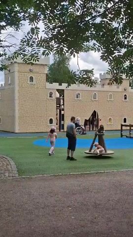 A drone shot pans across a playground on a cloudy day. The playground features a large, beige, castle-like play structure with turrets and windows. In front of the castle is a blue and green rubberized surface with various play equipment. A children's merry-go-round is visible with a child on it. To the right of the castle is a smaller, brown playhouse. People are scattered throughout the playground. A man is standing near the merry-go-round. A child is running near the castle. A person in a wheelchair is near the castle entrance. Two adults and two children are near the playhouse. A man is standing further away from the playhouse, holding something. Trees with green leaves frame the top of the shot. In the background, there are several wooden buildings with dark roofs. The ground in the foreground is covered in gravel.