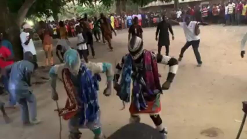 A crowd of people surrounds two costumed dancers in a dirt clearing. The dancers wear distinct masks; one has turquoise hair and the other a silver helmet-like mask. Both wear vibrantly colored clothing and perform animated, synchronized movements. The surrounding crowd watches and participates, with some clapping and dancing. The setting appears to be a rural or village environment with trees visible in the background. The lighting suggests daytime. The video is shot from a slightly elevated perspective.