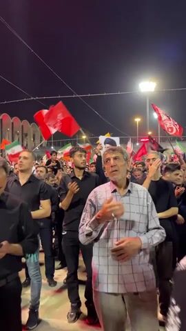 A nighttime outdoor gathering of people. Many individuals are holding red flags. One man in the foreground wears a checkered shirt and has his hand on his chest. Behind him, a young man in a black shirt also has his hand on his chest. Further back, a man is holding up two portraits of an elderly man with a beard and turban. Numerous other people are visible in the background, some also holding flags, including the Iranian flag. The lighting suggests streetlights or artificial illumination.