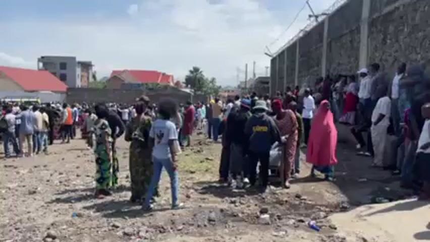 Here is a detailed description of the video:

**Overall Impression:** The video depicts a large crowd of people gathered in an open, possibly undeveloped, area. The setting appears to be outdoors, possibly in an urban or semi-urban environment. 

**People:**
*   There is a dense crowd of individuals of varying ages, genders, and ethnicities.
*   Clothing styles are diverse, ranging from casual wear like jeans and t-shirts to traditional garments. Some individuals are wearing head coverings.
*   The people are standing and milling about, appearing to be waiting or observing something.

**Objects:**
*   The ground is a mix of dirt, gravel, and scattered debris (possibly trash or small objects).
*   A tall concrete wall with barbed wire on top runs along the right side of the frame, possibly indicating a secure perimeter or property boundary.
*   Buildings with red roofs are visible in the background, suggesting a residential or commercial area.
*   A vehicle (possibly a car or van) can be seen in the left part of the frame.

**Actions & Interactions:**
*   The primary action is the gathering of people.
*   There appear to be minimal overt interactions or activities taking place within the crowd. People are mostly standing and observing.

**Environment:**
*   The weather appears to be sunny, with a clear sky visible.
*   The location seems to be on the edge of a developed area, given the presence of buildings and the somewhat undeveloped nature of the ground.

**Overall, the video portrays a crowd gathered in an open space, possibly for an event, demonstration, or other gathering. The surrounding environment suggests a location on the outskirts of a developed urban area.**