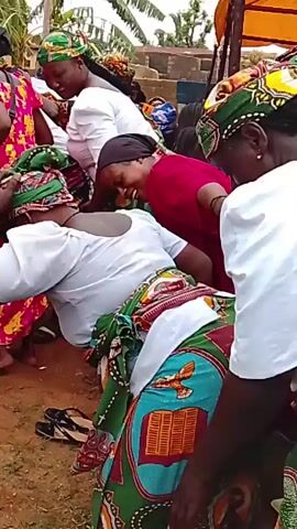 The video shows a group of women dancing outdoors, likely at a celebration or event. The women are wearing colorful traditional African clothing, including head wraps and patterned skirts or wraps. They are moving their bodies and swaying to the rhythm of music, although the audio in the provided excerpt is not very clear. The background shows a somewhat rural setting with some trees and a structure that appears to be a temporary tent or shelter. There are some children and other people visible in the periphery, suggesting a community gathering. The overall atmosphere seems lively and festive.