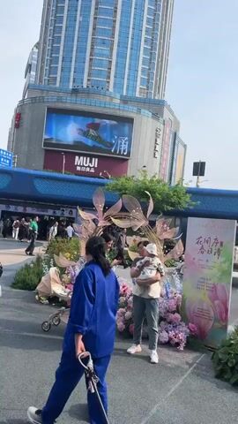 The video shows a public plaza in an urban setting. People are walking around, some are standing and talking, and others are taking pictures. There are several buildings visible, including a tall skyscraper with a MUJI store sign on it. There is a blue structure that appears to be an entrance to a subway or other transportation system. The plaza is decorated with flowers and plants. There is also a small kiosk.