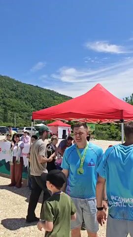 A man in a blue t-shirt and grey shorts with a medal around his neck looks forward, then right, as he walks towards a table under a red canopy. A small boy in a green t-shirt and black cap is to his left. A man in a grey t-shirt drinks from a white cup as a woman in a black tank top, black shorts, and a backpack pours a drink from a bottle into a small cup at the table. A small child with curly blonde hair stands next to the table, looking at the people around. The table is laden with many plastic water bottles. People in the background are standing and talking under the red canopy and other canopies. Mountains covered in green trees are visible in the background, and the ground is covered in pebbles.