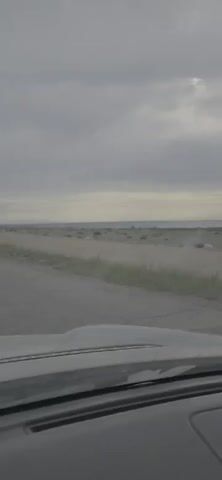 The video shows a view from inside a vehicle driving on a paved road. The road is cracked and appears to be in disrepair. To the left and right of the road is dry, grassy terrain with some scattered rocks. In the distance, there is a body of water, possibly the ocean, and a hazy horizon. The sky is overcast with heavy, gray clouds. The dashboard of the vehicle is visible in the foreground.