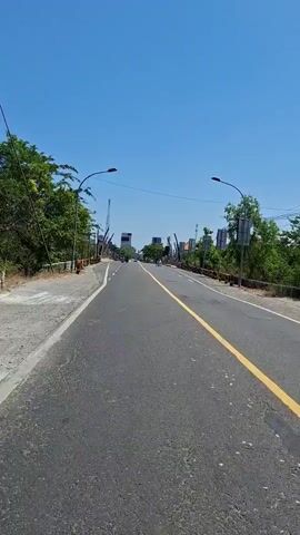 The video shows a view from a moving vehicle traveling on a paved road under a clear blue sky. The road is asphalt with a yellow line dividing the lanes. On both sides of the road, there are sidewalks, low stone walls with metal railings, and trees. Several lampposts line the road. In the distance, there are buildings and structures, including what appear to be construction cranes. Traffic is light, with a few motorcycles and a three-wheeled vehicle visible. The overall environment appears to be a sunny day in a semi-urban or suburban area.