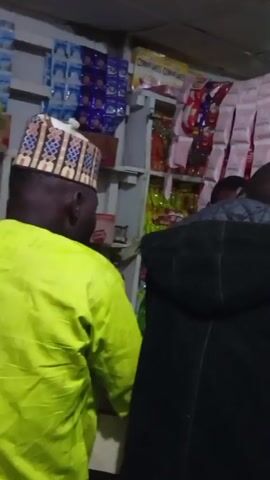 The video shows the inside of a small store or shop. Several people are present, browsing and presumably purchasing items. The store is stocked with various goods on shelves. The lighting is somewhat dim. The camera moves around, showing different individuals within the space. One person is seen using a mobile phone. Another person is wearing a red cap and blue shirt, carrying a bag.