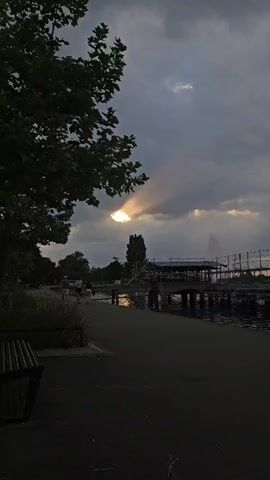 The video depicts a park scene on a cloudy day. Sunlight breaks through the clouds, casting a ray on the water. A walkway runs along the water's edge, with a bench to the left. In the distance, there is a fountain in the water and a structure, possibly a bridge, under construction with scaffolding surrounding it. People can be seen walking along the path in the background. A tree frames the left side of the shot. The scene appears calm and somewhat subdued in lighting.
