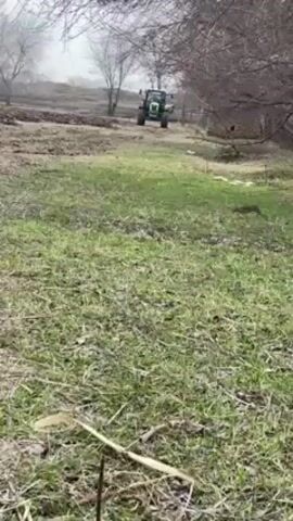 The video shows a green tractor in a grassy field on a foggy day. The tractor is positioned on the far side of the field, facing the camera. Some bare trees are present on both sides of the field.  The grass is green with some dead leaves scattered throughout. The tractor appears stationary.