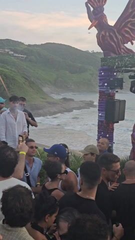 The video shows a crowd of people at an outdoor music event. A DJ is performing on a stage that is decorated with colorful sculptures, including a large bird-like figure and a tentacle-like structure. The event appears to be taking place on a beach or near the ocean, with hills and trees visible in the background. The lighting is bright and colorful, and the crowd seems to be enjoying the music and atmosphere. Many attendees are filming the show with their phones. The sky is overcast.