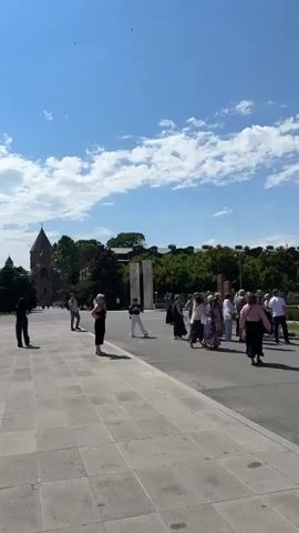 The video shows a panning shot of a large, open, paved square in front of several buildings. The sky is bright blue with scattered white clouds. In the foreground, the paved square is made of large, light-colored square tiles. In the middle ground, there are a few people standing around and walking in the square. There is a large, leafy tree on the left side of the frame, casting a shadow on the ground. In the background, there are several buildings, including a tall, dark tower with a spire on the left side, a long, light-colored building with arched windows in the center, and other structures. People are walking on the paved areas and on grass in front of the buildings. The overall impression is of a public space on a sunny day.