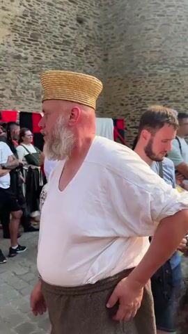 A man wearing a straw hat and a white tunic stands in the foreground, facing left. Behind him, a crowd of people gathers in front of a stone tower and tents. Two men in historical costumes, holding spears, appear to be part of a reenactment or performance. The sun shines brightly, casting lens flares into the scene.
