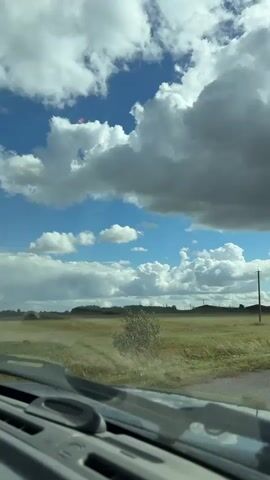 The video shows a view through a vehicle's windshield looking out at a rural landscape. The sky is filled with large, white and gray clouds against a bright blue background. The ground is covered in dry grass, with some faint tracks visible. In the distance, there are trees and some structures, possibly buildings or power lines. A single utility pole stands on the right side of the frame. The windshield wipers are visible in the foreground, indicating the vehicle is in motion. The overall scene suggests a cloudy day in an open, possibly agricultural area.