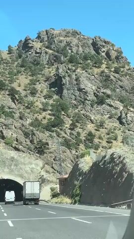 The video shows a view from inside a vehicle traveling on a highway towards a tunnel entrance. The tunnel is carved into a rocky mountainside. A white van and a white truck are visible in front of the vehicle, both heading into the tunnel. The tunnel entrance is illuminated, and the mountainside is covered with sparse vegetation and large rock formations. The sky is clear and blue.