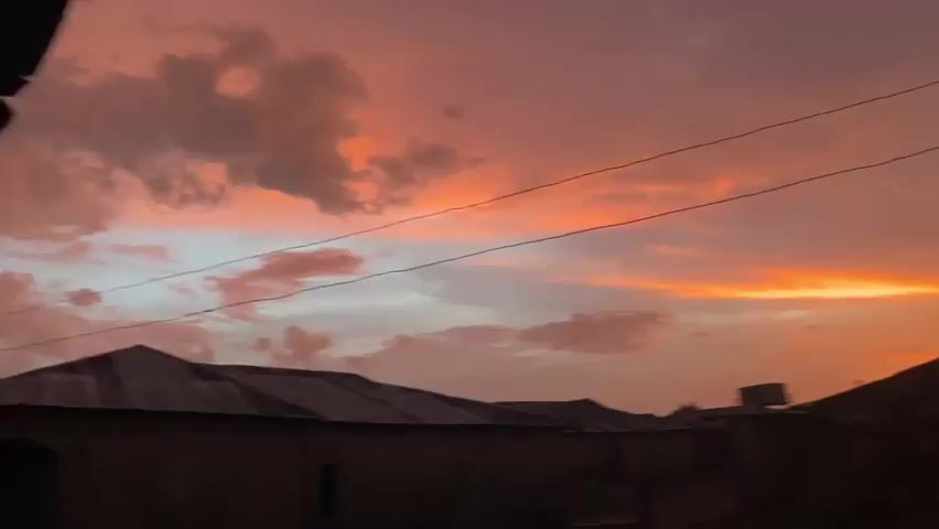 The video shows a vivid sunset with orange and pink hues in the sky, partially obscured by dark clouds. Silhouetted rooftops of buildings are visible at the bottom of the frame. Two power lines stretch across the sky. The camera pans upwards slightly, revealing more of the sky.