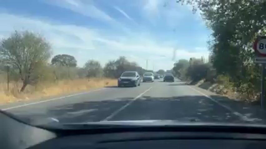 A vehicle is being driven on a two-lane road with a white line separating the directions. Other cars are on the road, traveling in the same direction as the vehicle with the camera. The sky is blue with scattered clouds. Bushes and trees line the side of the road.