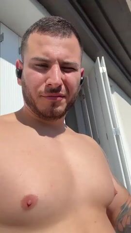 A medium shot depicts a shirtless man smoking a cigarette on a balcony in the sunshine. He has short dark hair, a beard, and a tattoo on his right arm. He is wearing wireless earbuds. The balcony railing is white and a white wall with vertical slats is partially visible behind him. The camera is held at chest height.
