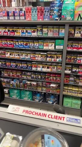 The video shows a shelf in a store stocked with packs of cigarettes and other tobacco products. The camera pans across the shelves, showing various brands and types of products. The products are arranged in rows and columns, with some products in boxes and others in individual packs. The lighting in the store appears to be fluorescent. The video also shows a sign that says "PROCHAIN CLIENT" which means "NEXT CUSTOMER" in French, indicating the location is likely in a French-speaking region. There are no people visible in the video.
