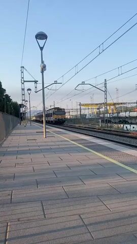 The video shows a train arriving at a station. The train is visible in the distance and is moving towards the platform. The platform is paved with grey tiles and has yellow safety lines along the edge. There are several lampposts on the platform. In the background, there are train tracks, overhead wires for the electric train, and some industrial buildings. The sky is clear and blue, indicating it is daytime. The sun is casting long shadows on the platform, suggesting it is either early morning or late afternoon. The train appears to be a passenger train. There are no people clearly visible on the platform or in the train. The overall scene is one of a quiet train station during daylight hours.