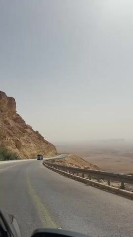 The video depicts a desert road with a truck driving towards the camera. The road is asphalt with yellow lines and metal guardrails on the right side. To the left is a rocky hill. Beyond the guardrail is a desert landscape with a hazy sky. The video is shot from inside a car, with a portion of the dashboard visible.