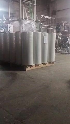 The video shows a pallet of large white rolls wrapped in plastic inside a dimly lit industrial warehouse. In the background, a large piece of machinery with lights is visible, possibly a production machine. The floor is concrete and appears dusty. The rolls are stacked upright on a wooden pallet. The lighting is artificial and harsh, casting shadows. The overall impression is a storage area within a factory or processing plant.