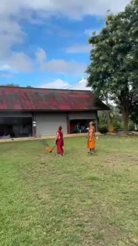 Two people dressed in traditional Buddhist monk robes stand on a grassy field near a building with a red roof. The person on the left is sweeping the grass, while the person on the right is observing the sky. The camera pans up to reveal a small, decorative hot air balloon ascending into a cloudy sky. Smoke trails behind the balloon as it rises.