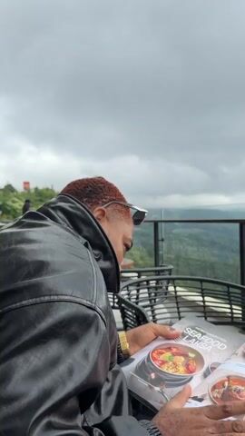 A person wearing a black leather jacket with red hair is seated at an outdoor table, looking at a menu. The table is made of wood and has a glass railing behind it. The railing overlooks a lush green mountain landscape under a cloudy sky. The menu displays images of food, including "SEAFOOD LAKSA". A cell phone and a cigarette pack are also visible on the table. The camera pans across the table and the scenic view.