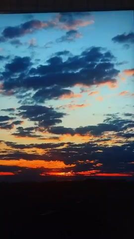 The video shows a dynamic sunset with clouds. The sky transitions from a deep blue at the top to orange and red hues near the horizon. Dark clouds are scattered throughout, some tinged with pink and orange from the setting sun. The bottom of the frame is obscured by a dark silhouette, likely indicating land or a landscape feature. The overall impression is one of natural beauty and the changing light of day.