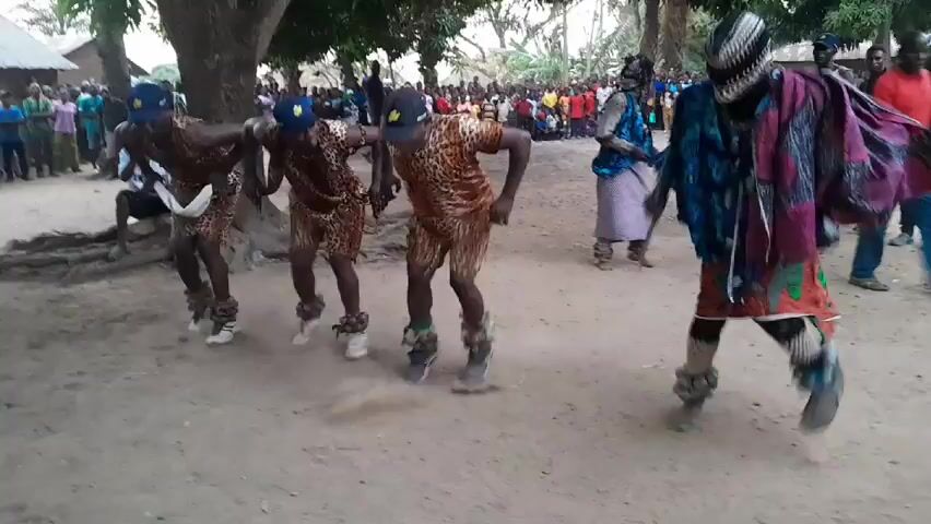The video shows a group of four men dancing in a dirt clearing in front of a crowd of people. The dancers are wearing leopard-print outfits with caps and bell-like adornments around their ankles. The dancer to the right of the group wears more colorful, layered clothing and carries a stick. The background shows trees and simple structures. The camera appears to be held by someone within the crowd, recording the dance.