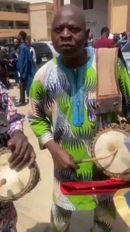 Two men wearing traditional African clothing are playing drums outside a building. The man on the left is wearing a white and red patterned shirt and a striped hat, while the man on the right is wearing a green and blue patterned outfit. Each man has a drum strapped to his body and is playing it with his hands and a small wooden stick. There are other people and cars in the background. The scene appears to be taking place during the day.