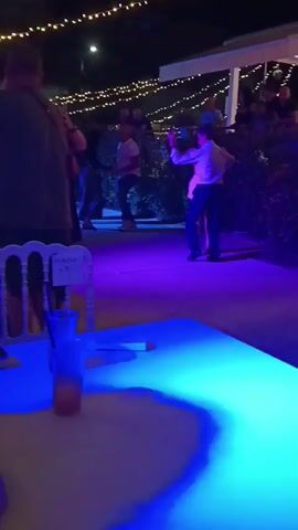 The video shows a group of people dancing outdoors at night. There are string lights overhead, and the area is lit by colored spotlights, primarily blue and purple. In the foreground, a table with a drink and a phone is visible. The main action features several couples dancing. One couple in the center is dancing closely, with the woman in a white top and dark pants and the man in a dark shirt. To their right, another couple is dancing, with the woman in a white skirt and the man in a light-colored shirt and dark pants. Further to the right, another couple is dancing, with the woman wearing a patterned top and dark pants. Other people are visible in the background, some watching and some dancing. The scene appears to be a social gathering or event.