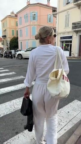 A woman is walking across a crosswalk. She has blonde hair styled in a bun. She is wearing a white outfit consisting of a long-sleeved shirt and pants, and carrying a white tote bag over her shoulder and a black item in her hand. She is also wearing sunglasses and hoop earrings. The background includes buildings with pastel colors and blue shutters, parked cars, and motorcycles. The sky is slightly cloudy. The scene appears to be a street in a European town.